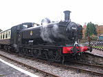 A black pannier tank locomotive is passing along a single track through woodland. The locomotive is pulling a train of a covered red wagon, three grey open wagons, and a red guard's van.