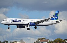 A white plane with the words "jetBlue" painted at the front and a blue-green tailfin approaches landing with its landing gear deployed as it soars above houses below