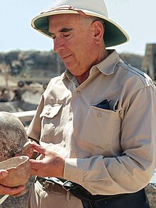Photograph of an elderly man holding an ancient Greek pot, wearing a khaki suit and pith helmet.