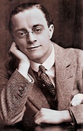 Black and white photo of a bespectacled man in suit and tie resting his elbows on a table, with his face resting against his right hand