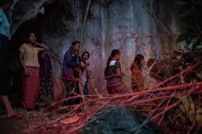 Women and children taking shelter in a cave from an air strike in Karenni State. It's quite dark, There are branches piled up in front of the area where they are walking.