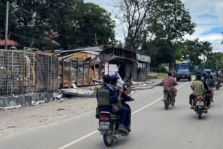 Two members of the MNDAA police on a motorbike in Lashio. There are a few other people on the street. Buildings on their left look like they've been burned down.