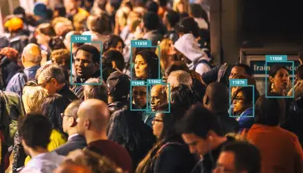 A crowded subway platform shows several commuters turning to face the viewer. Their heads are surrounded by digital boxes that label each an arbitrary number, implying categorization by facial recognition software. The commuters appear to be of various races and ages.