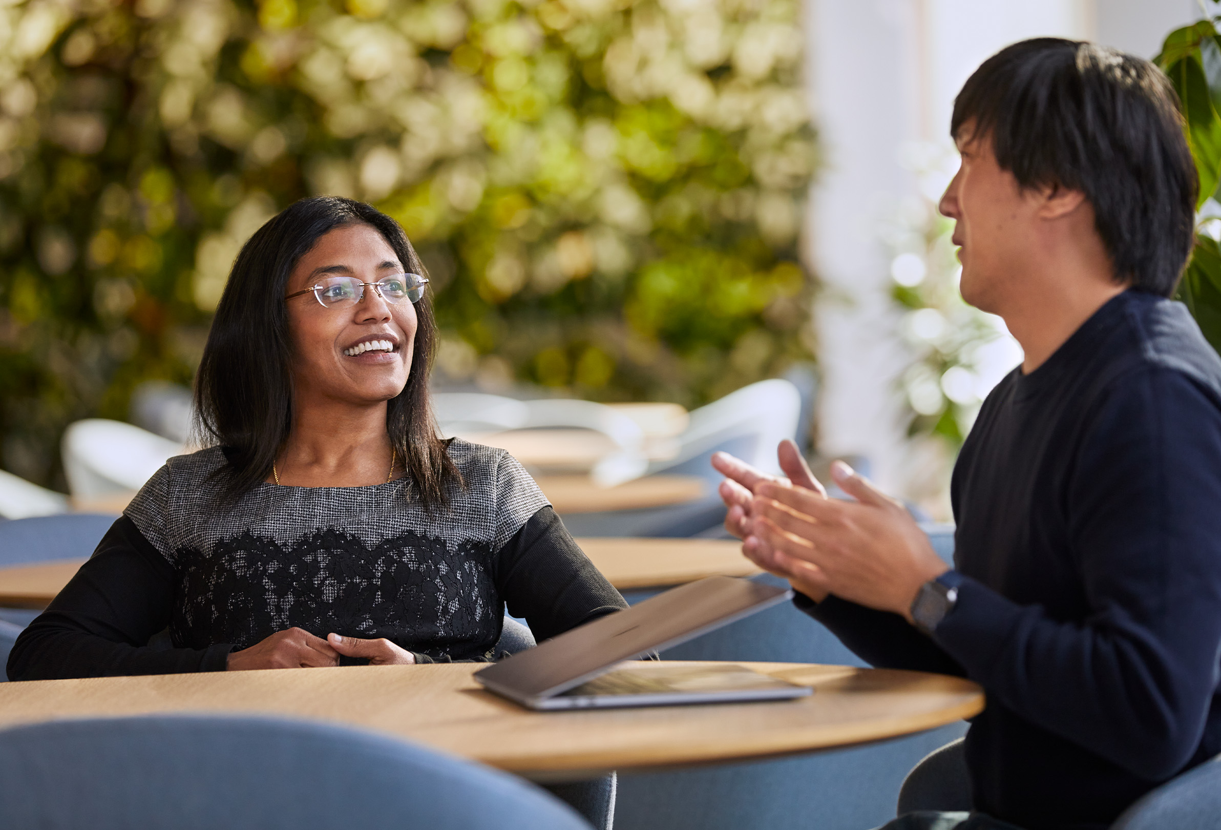 Two smiling Apple employees sitting outside with a MacBook and greenery in the background.