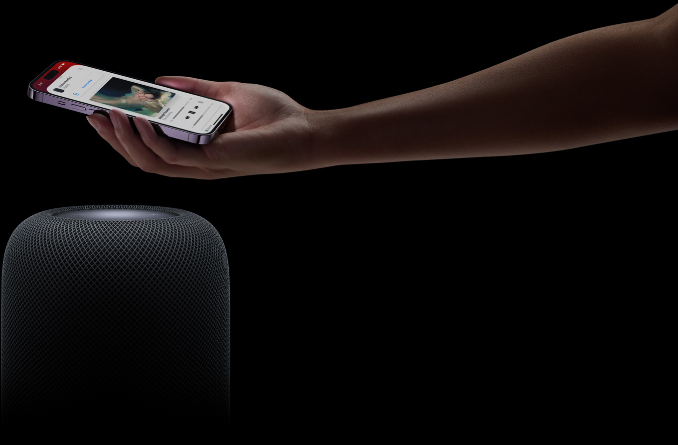 มือข้างหนึ่งยื่นมาจากทางขวาของเว็บเพจโดยที่ในมือถือ iPhone อยู่เหนือลำโพง HomePod