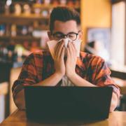 Person covering their face with a tissue while working on a laptop