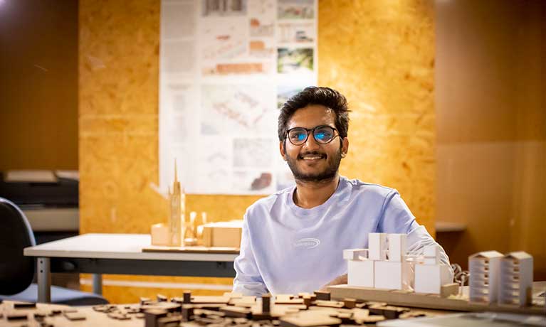 Male student smiling at camera with architectural models on the table in front of him.