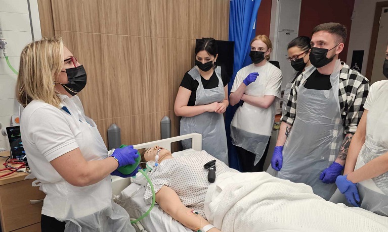 A group of people wearing face masks, aprons and gloves stood around a hospital bed containing a dummy patient