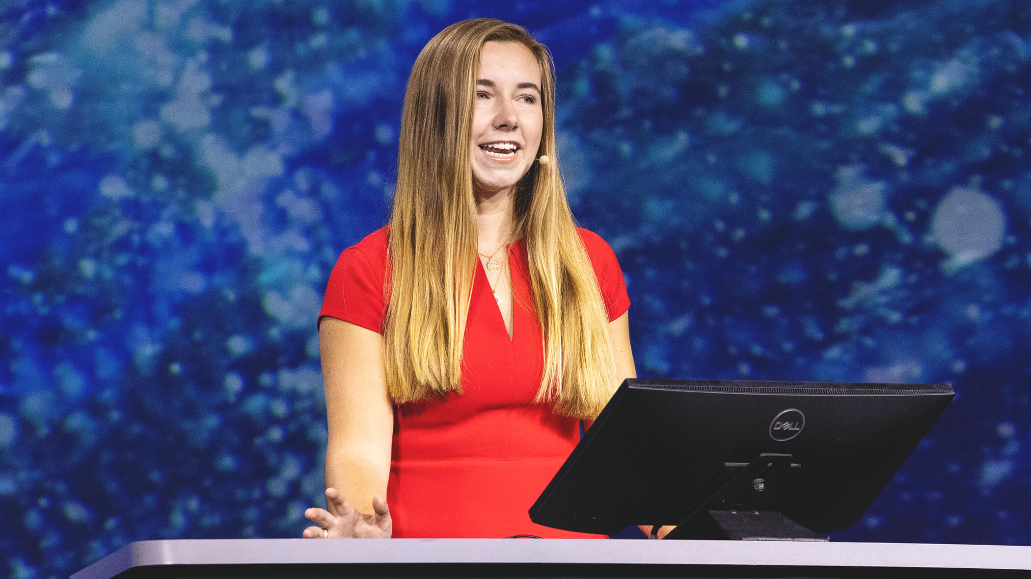 Presenter with long straight blonde hair, wearing a red top, speaks from the stage against a blurred blue background