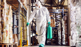 Image of man in protective suit and mask disinfecting warehouse full of food products