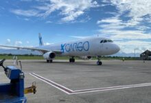 A GlobalX cargo jet in light blue lettering on tarmac of small tropical airport as seen from ground level in front, blue skies above.