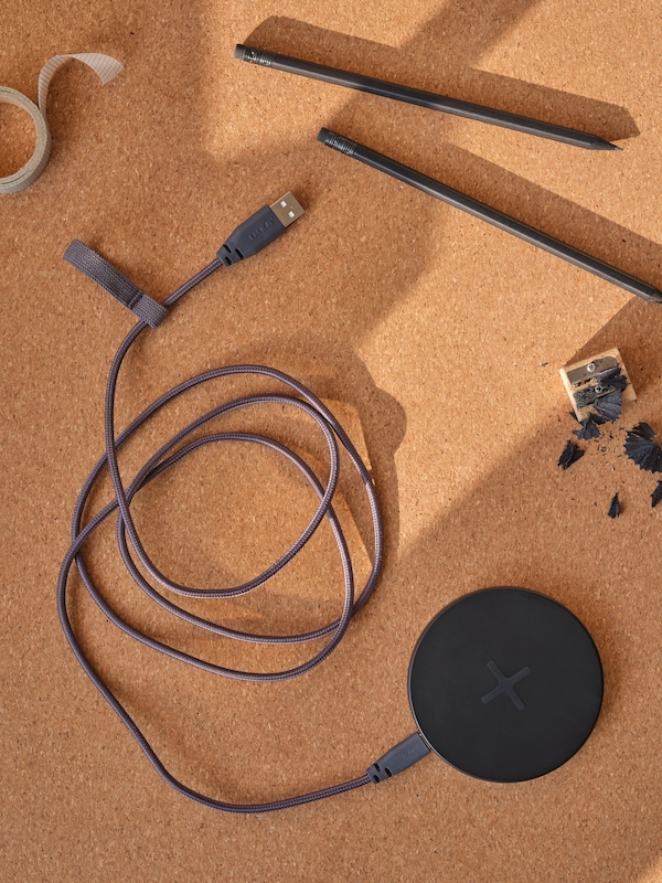 A wireless charger cable sitting on top of a corkboard with two pencils lying beside it