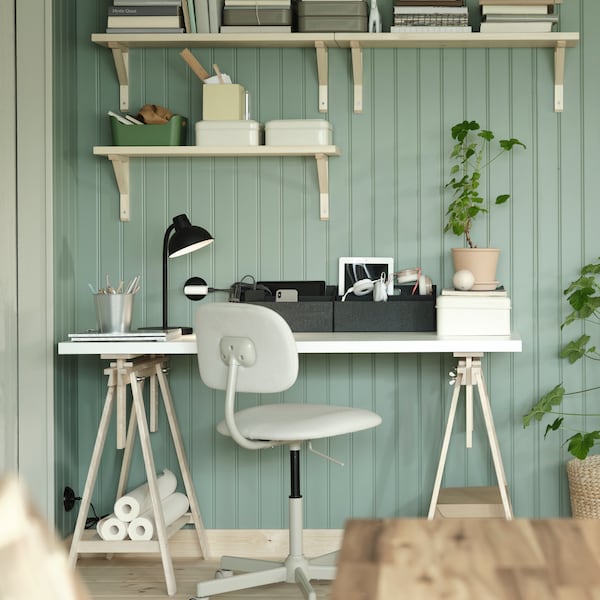 A workstation placed against a green wood-paneled wall with wall-mounted TRANHULT shelves and a BLECKBERGET swivel chair.