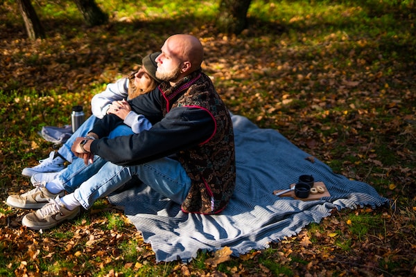 A man and a woman sitting on a picnic blanket in the woods covered by dried autumn leaves.