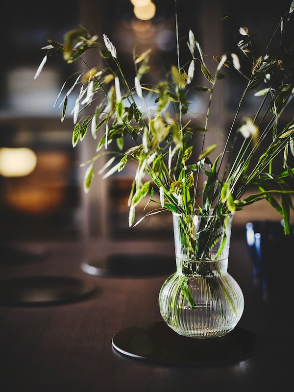 A PÅDRAG glass vase with green foliage sits on a dining table on a black round placemat. More placemats are dotted around.