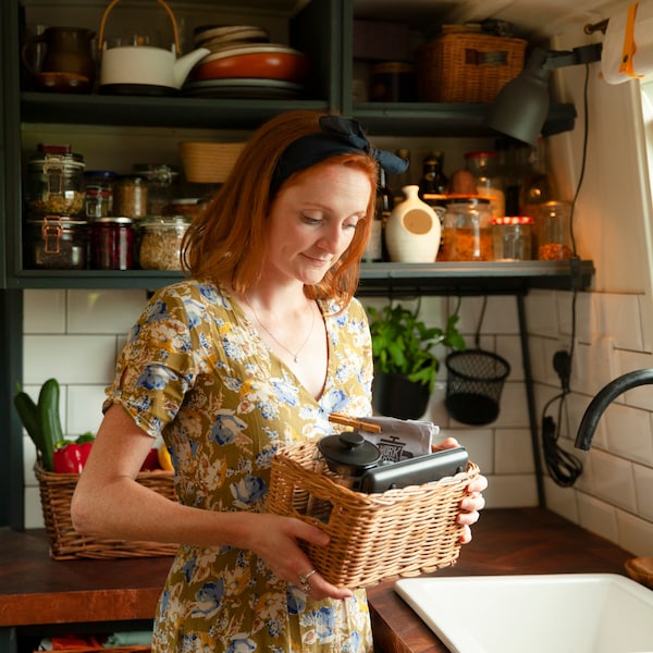 A woman in a kitchen carries a small basket, containing her PÅTÅR coffee, UPHETTA coffee maker and GENOMSITT electric scale.