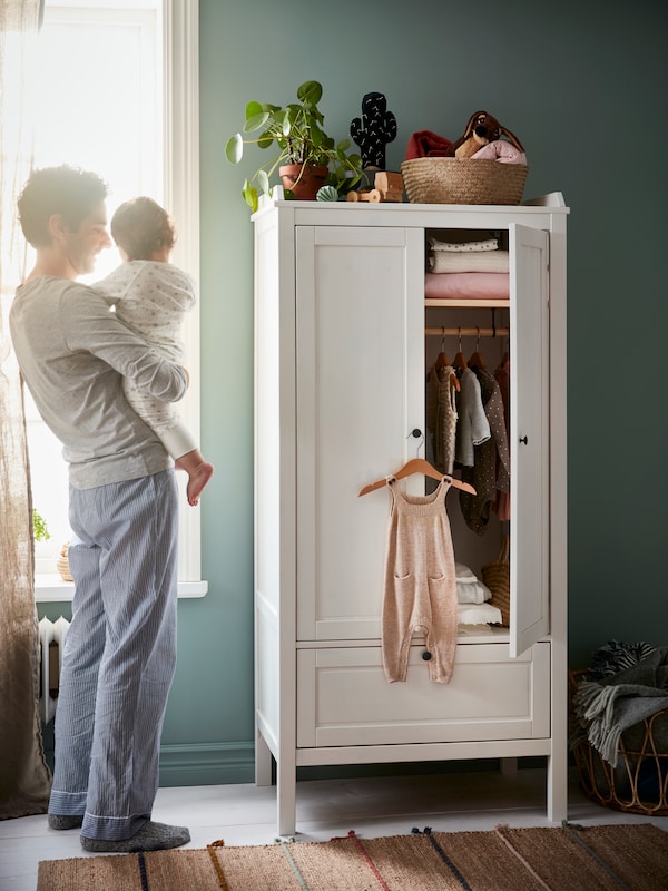 A white SUNDVIK wardrobe with clothes hanging in it and a man holding a baby smiling next to it.