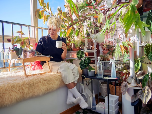 Man sitting next to a window, reading a book and drinking coffee, surrounded by house plants.