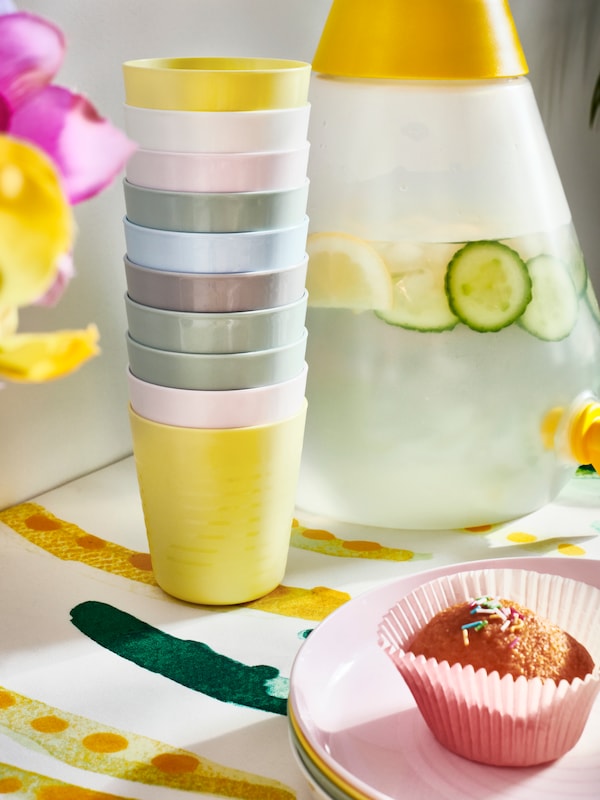 Colorful kids cups and plates next to a jug of water with cucumbers and lemons in it.