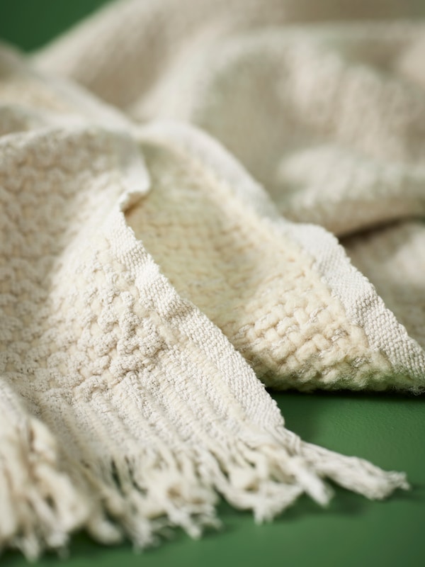 The detailing, weave, and fringes of a chunky LAPPTÅG throw in off-white against a green backdrop.