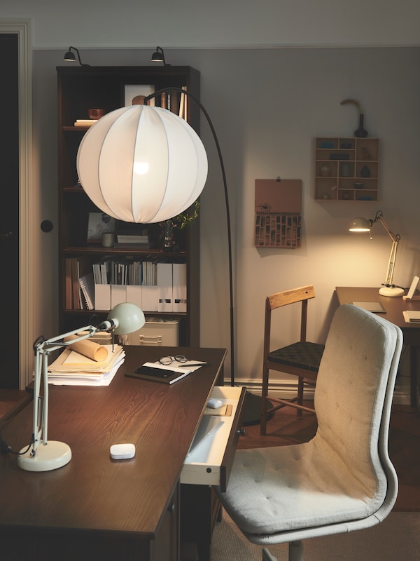 A refined workspace at home has an arched floor lamp hanging over an IDANÄS desk with a drawer open, and a work lamp on it.