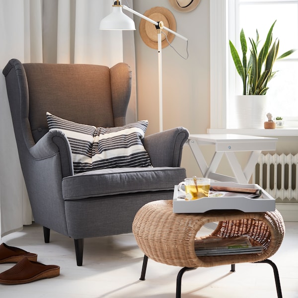 A white RANARP floor/reading lamp placed behind a grey armchair in the corner of a bright living room with beige walls.