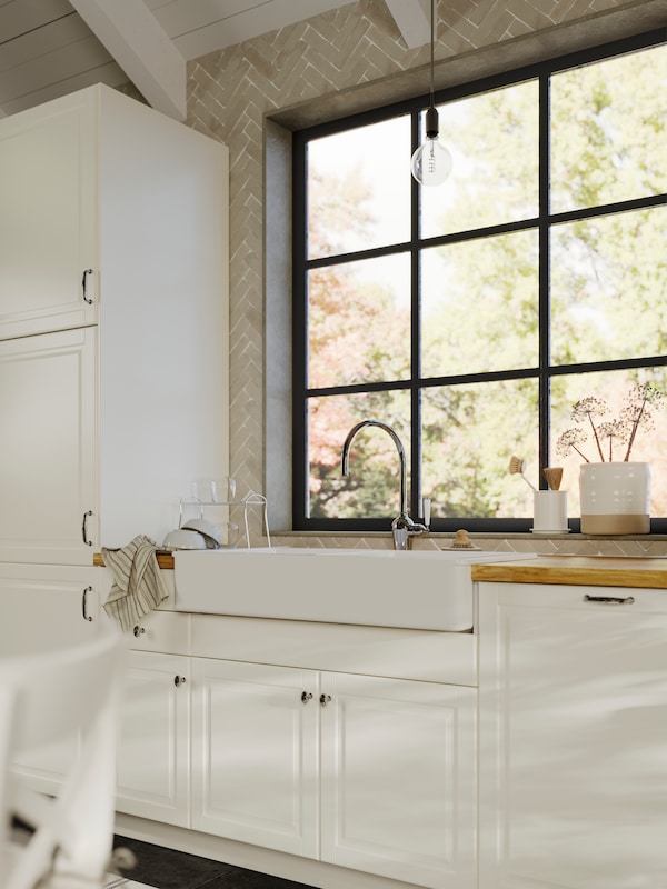 A kitchen in traditional style with BODBYN fronts in off-white and HAVSEN sink bowl, by a large industrial-styled window.