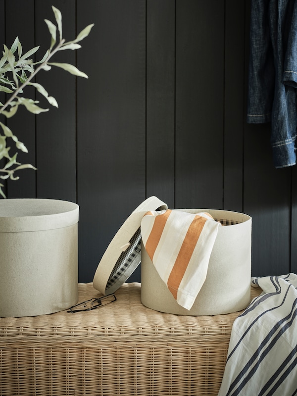 Two small round decoration boxes with lid and white on a rattan bench. Striped towels in blue and orange around it.