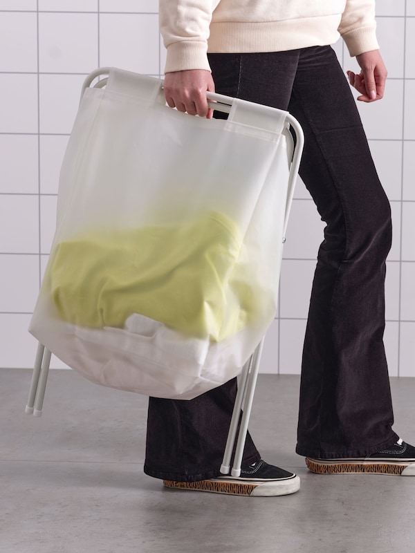 A person carrying a folded JÄLL laundry bag with stand with some laundry in it.