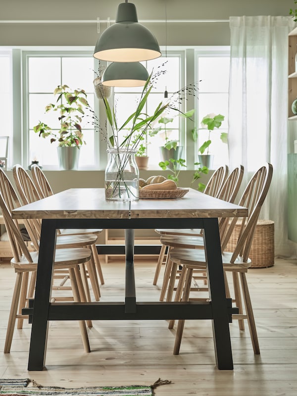 In a light-filled dining room are a large SKOGSTA table and six chairs in solid acacia, a glass vase with wild flowers.