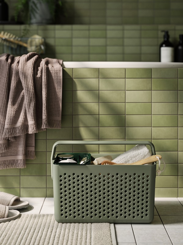 A gray-green UPPRÄMEN storage basket filled with body care items is standing on the bathroom floor in front of a green bathtub.