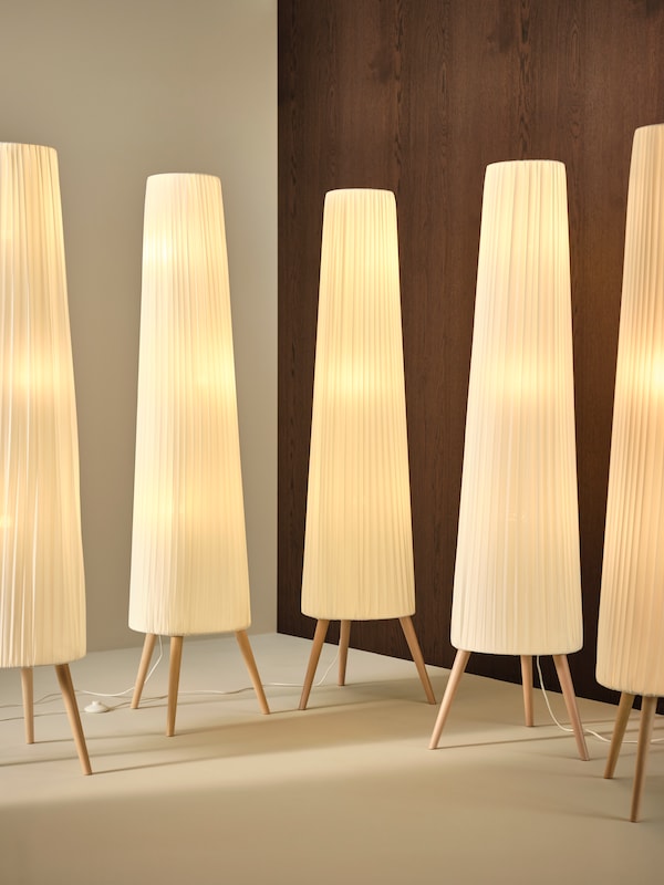 A neatly arranged row of lit ÖKENSAND floor lamps, in a corner of a room, against a cream-coloured wall and a dark wood wall.