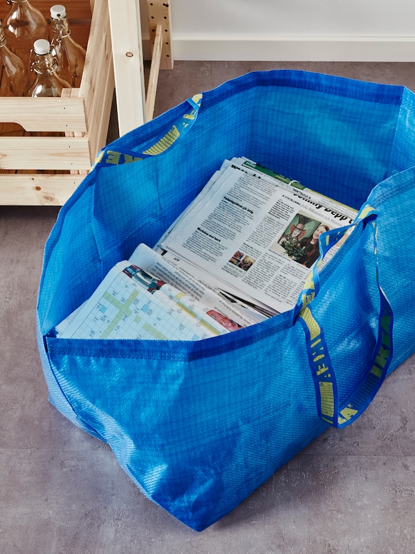 A blue FRAKTA bag, half full of newspapers, on the floor in front of a box of clear-glass KORKEN bottles with stopper.