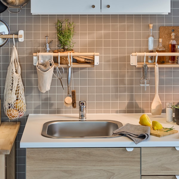 A white/oak effect ENHET kitchen by a grey-tiled wall with a birch rail, two birch wall shelves and a white wall cabinet.