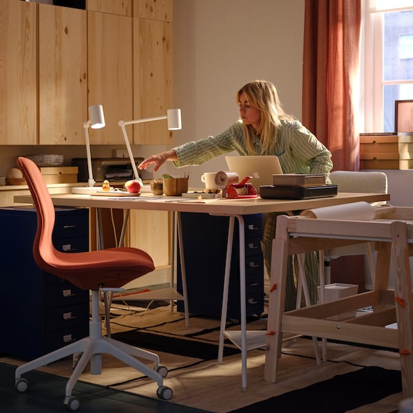 A workspace with IVAR cabinets and a LÅNGFJÄLL office chair. A person sitting behind a desk while reaching for an apple.
