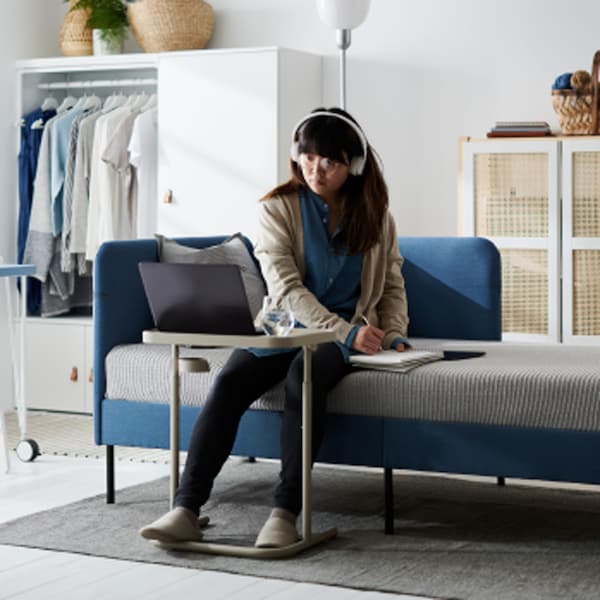 A young woman with headphones sits on the side of a BLÅKULLEN bed, focusing on a laptop placed on a BJÖRKÅSEN laptop stand.