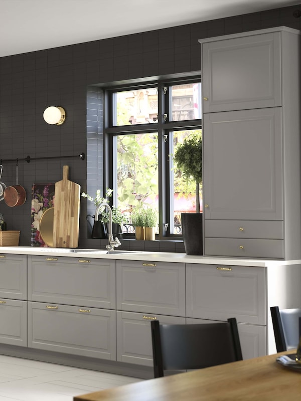 An organized gray BODBYN kitchen with a cutting board sitting on the countertop and a big window above the sink.