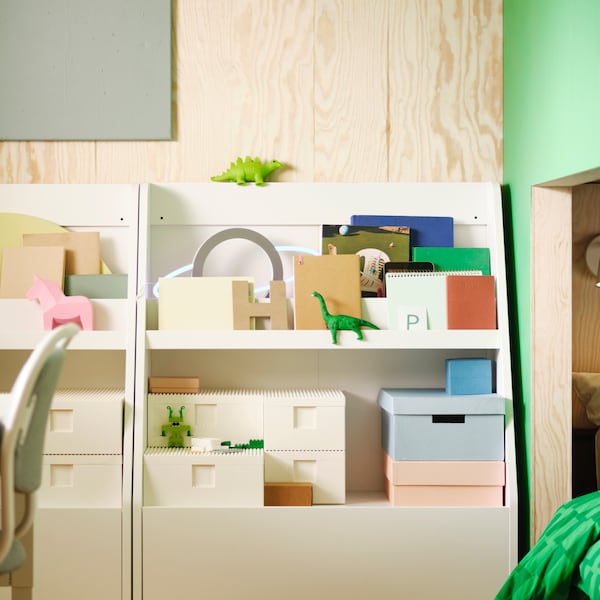 Two white BERGIG book displays with storage, holding a variety of boxes, books and toys, stand against a wall.