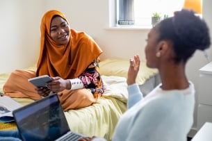 Two female university student friends in their dorm bedroom are studying together while sitting on the bed