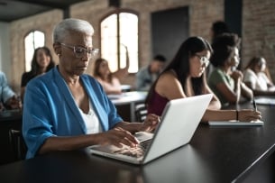 A senior woman with white hair types on her open laptop in a classroom with younger students in the background.