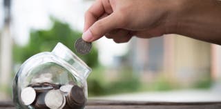 A hand dropping a coin into a savings jar that has other coins