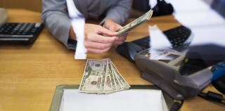 A person holding a stack of money with a calculator and computer beside them, while a receipt is flying out of a printer 