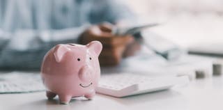 A pink piggy bank on a desk in the foreground with a person sitting at the desk in the background.