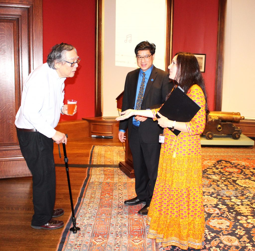 Color photograph of three people in a wood-paneled room with art on the walls and a cannon in the background, with a decorative rug on the wooden floor. There are two men, one older, one middle-aged, and a middle-aged woman, all three are Asian of different backgrounds. The man to the left is wearing a white shirt with black pants and leans onto a cane while listening and holding a beverage. The man in the middle is wearing a black suit jacket and pants with a blue collared shirt and multicolored tie. The woman on the right is wearing a yellow dress with pink filigree and is holding a black clipboard while speaking and gesturing to the man on the left. 