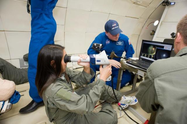A woman sits on the floor and looks through an eyepiece device. She is wearing a light brown flight suit. Two people are behind her in blue flight suits and one person in front of her to the right is wearing a light brown flight suit.