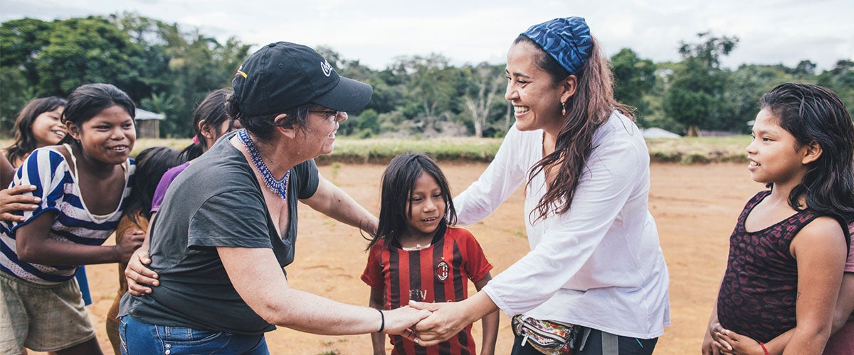 Two woman embracing a young girl in an open field surrounded by a group of children. 