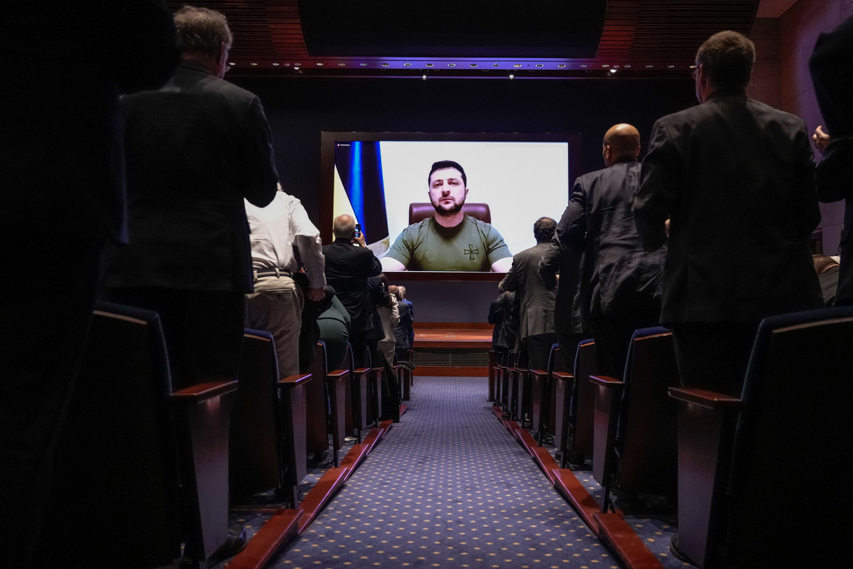 Ukrainian President Volodymyr Zelenskyy appearing on a large screen, while people in the audience stand.