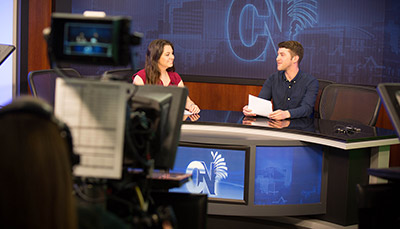 journalism students sat at news desk with student and camera in foreground