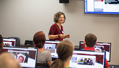 Teacher in front of class room with students sat behind computers.