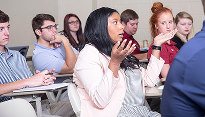 Student in a desk talking out loud in a classroom with other students. 